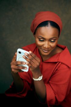 Portrait of a smiling woman wearing a red headscarf, holding a smartphone indoors.