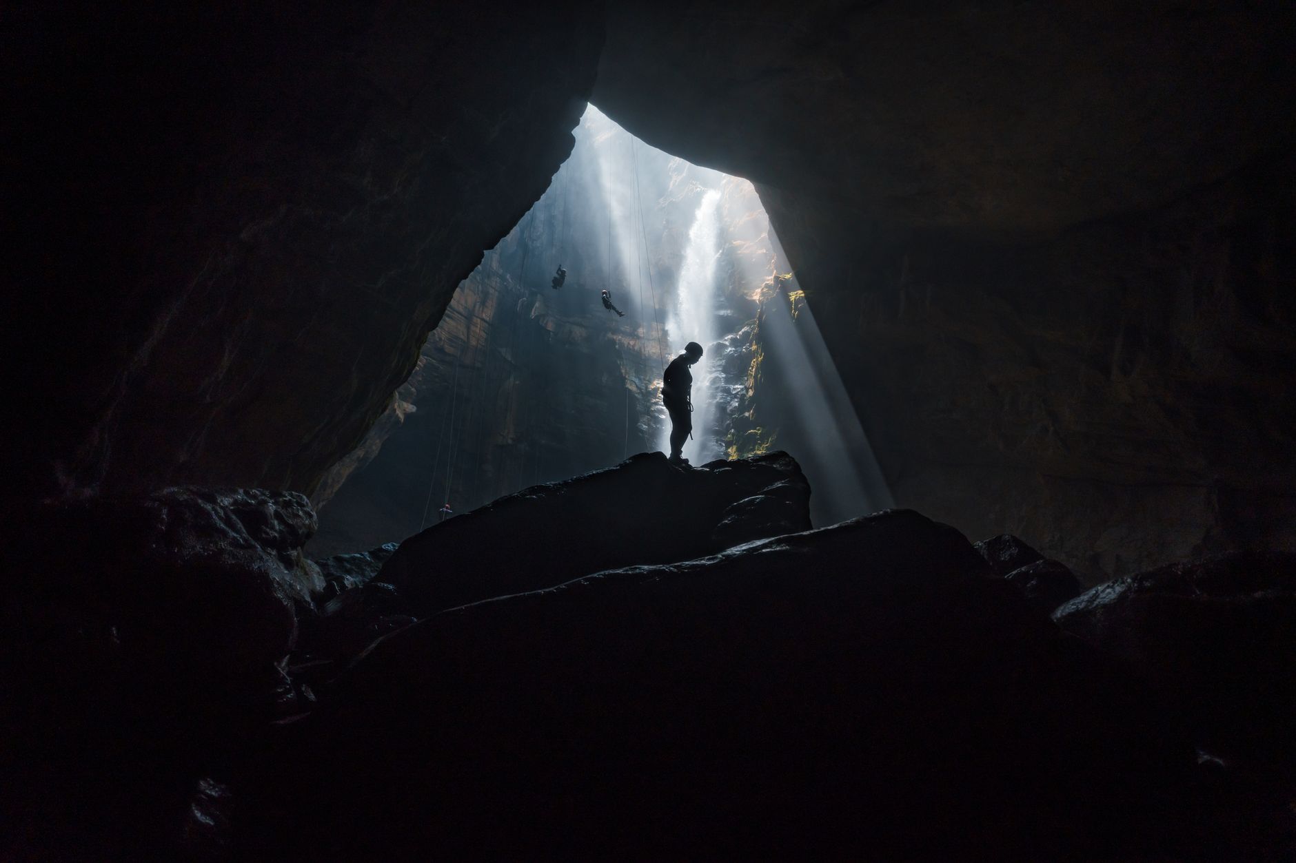 Silhouette of a person exploring a vast cave with striking sunbeams filtering through the opening.