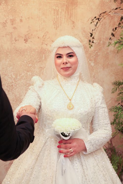 Free Bride in ornate white gown holding a bouquet, hand in hand indoors. Stock Photo