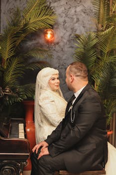 Bride and groom seated by a piano, enveloped in lush greenery, gaze lovingly at each other.