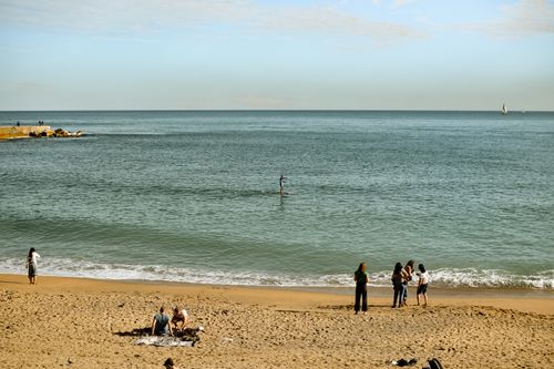 Free A diverse group of people relax and enjoy leisure time on a sunny beach, with a paddleboarder in the distance. Stock Photo