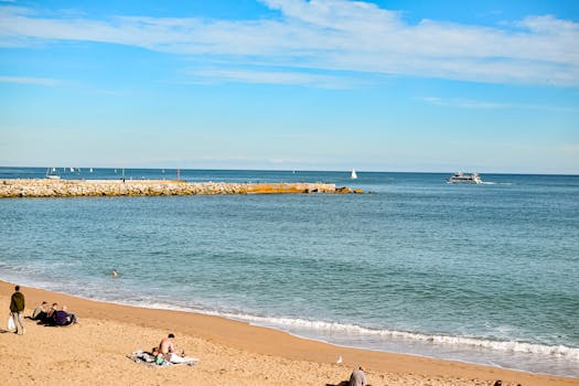 A lively beach scene with people relaxing, boats sailing, and a clear blue sky, evoking a sense of summer tranquility.