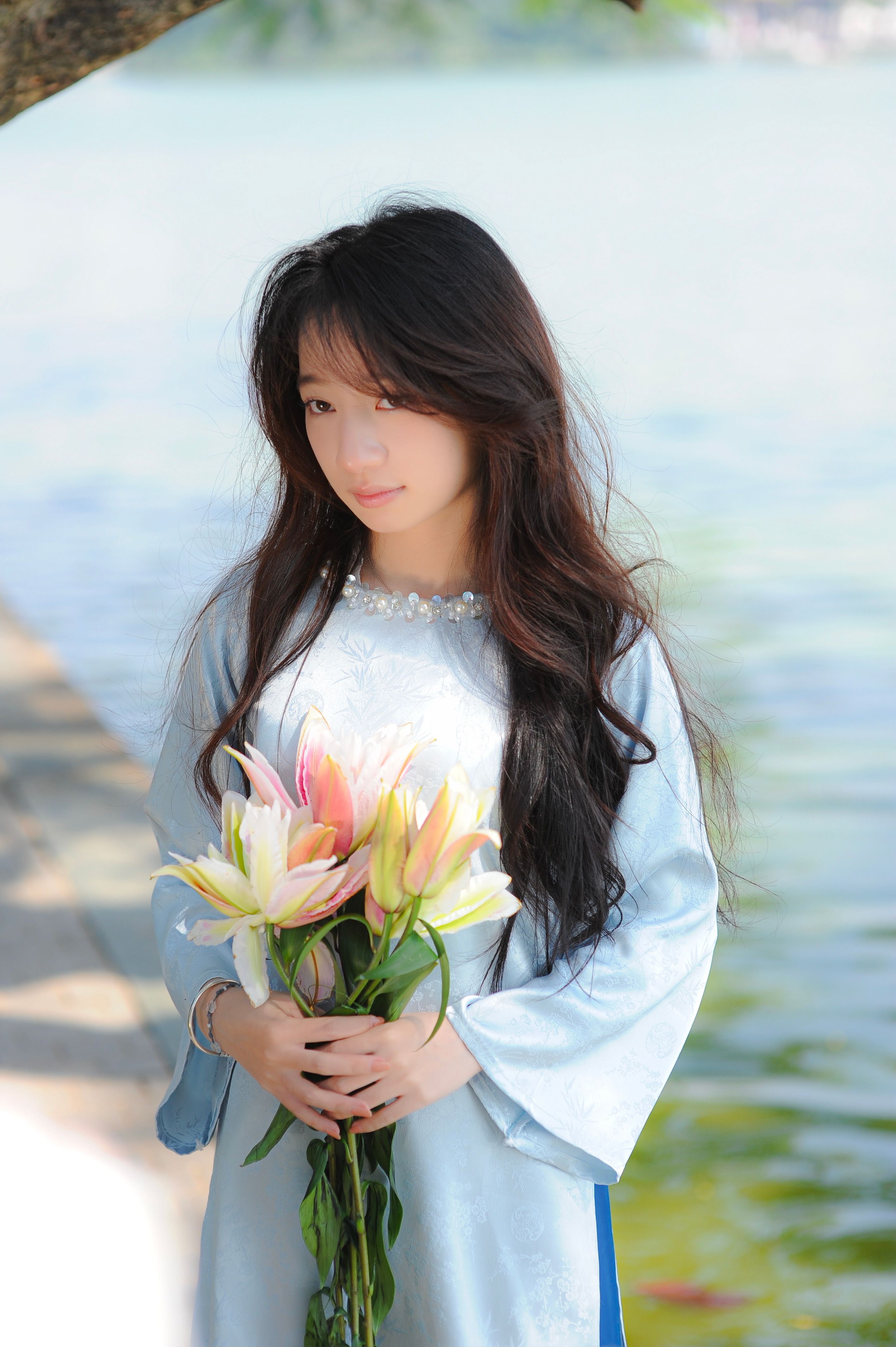 Elegant woman in traditional dress holding lilies near water on a sunny day.
