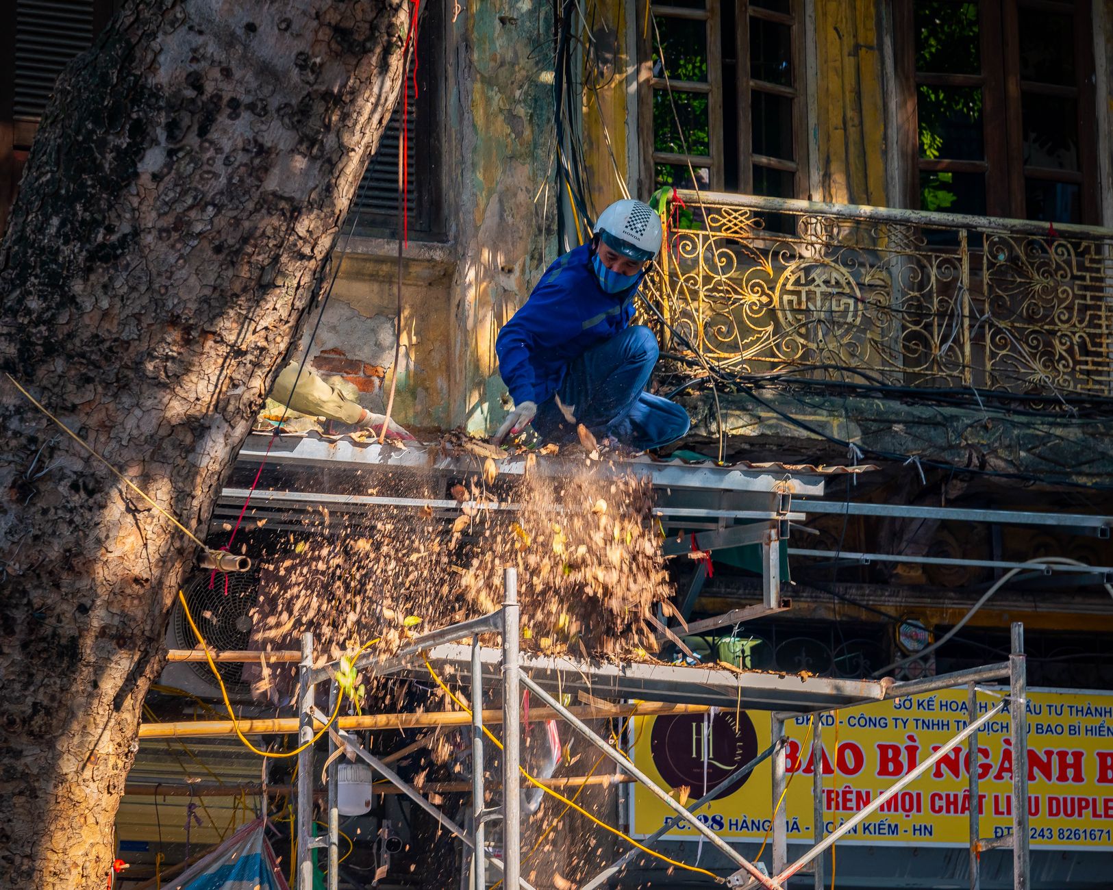 A construction worker in safety gear handling equipment on a scaffold. Dust and debris are visible.