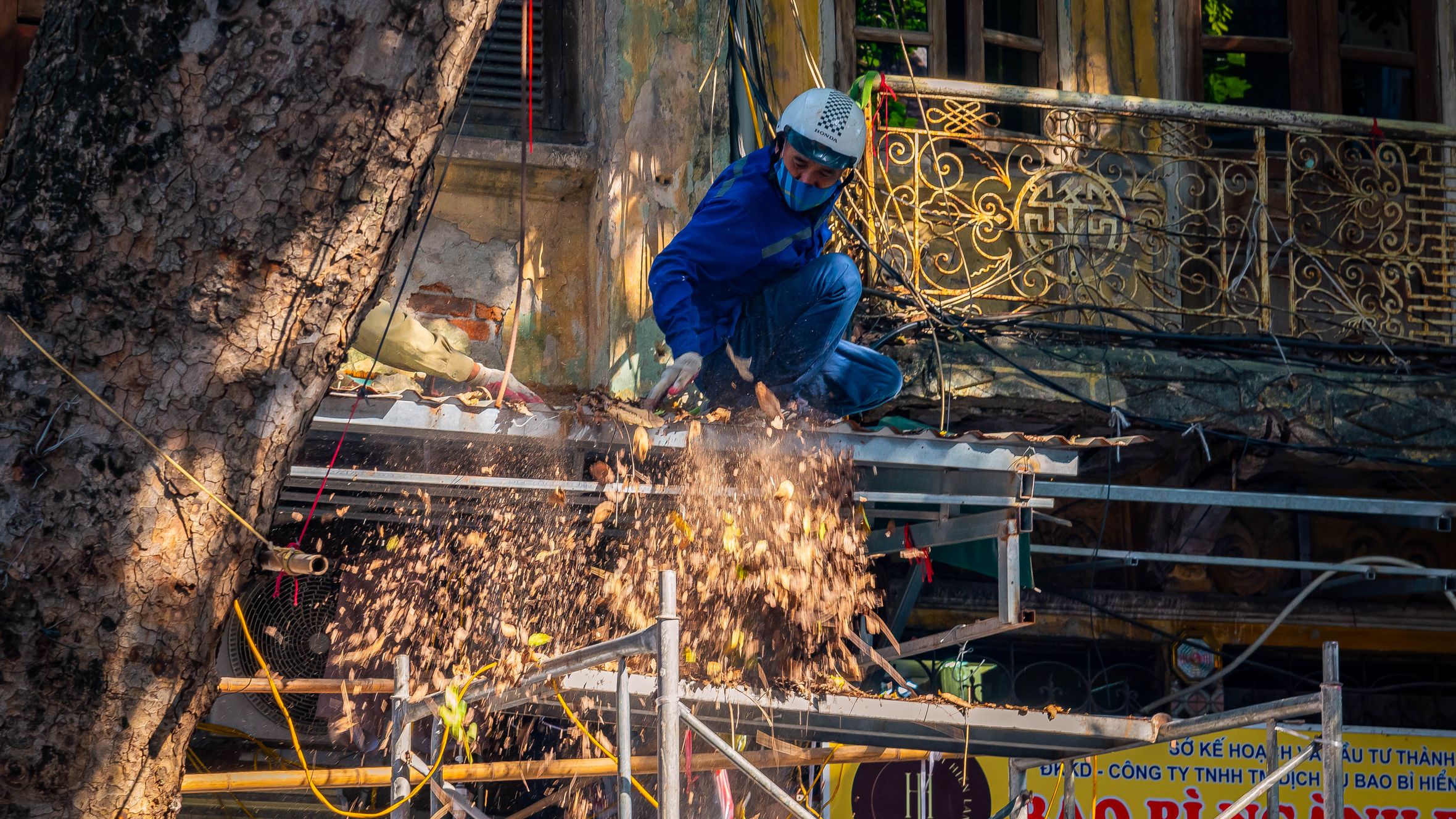 A construction worker in safety gear handling equipment on a scaffold. Dust and debris are visible.