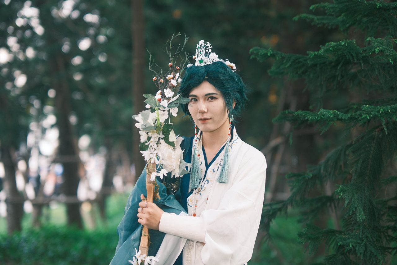 a person in elaborate cosplay attire holds a floral staff in a forest