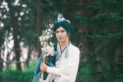 Cosplayer in traditional attire holding a floral staff, set against a forest backdrop.