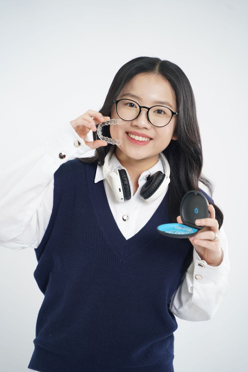 A teenage girl in a school uniform smiling while holding a clear Invisalign aligner and its case, representing Invisalign for teens in Bloor West Village, Toronto