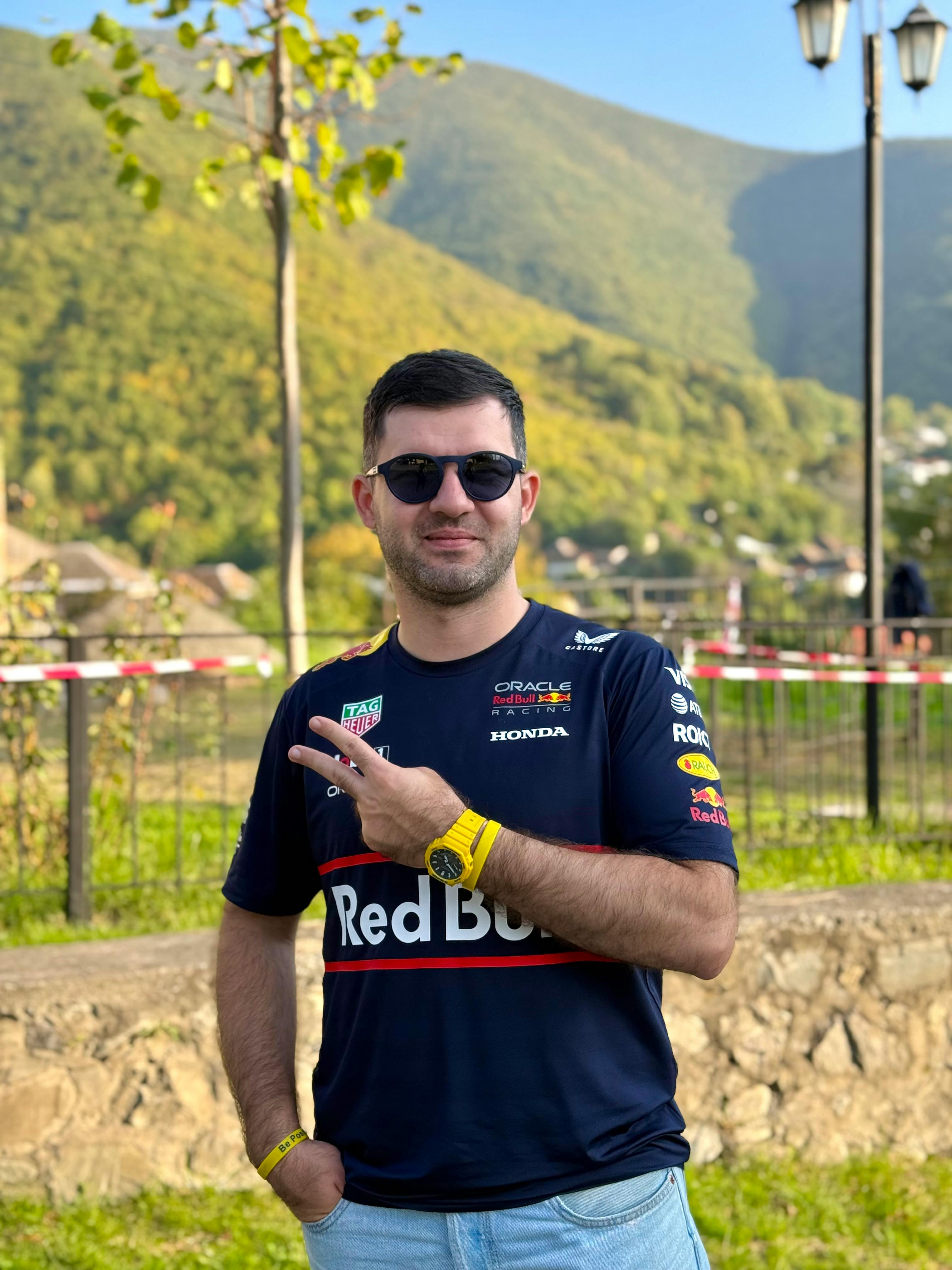 Smiling man in sunglasses and Red Bull shirt showing peace sign outdoors in scenic valley.