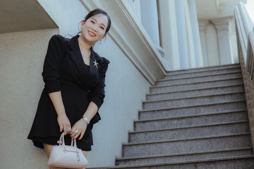 Young woman in stylish black attire smiling and holding a handbag beside a staircase.