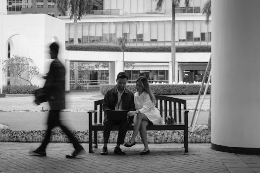 A couple using a laptop on a bench outdoors in an urban environment, with a passerby.