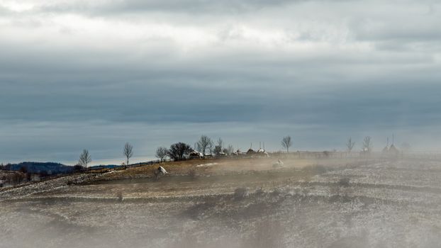 Rustic winter landscape featuring misty hills, haystacks, and a quiet village under a dramatic sky.