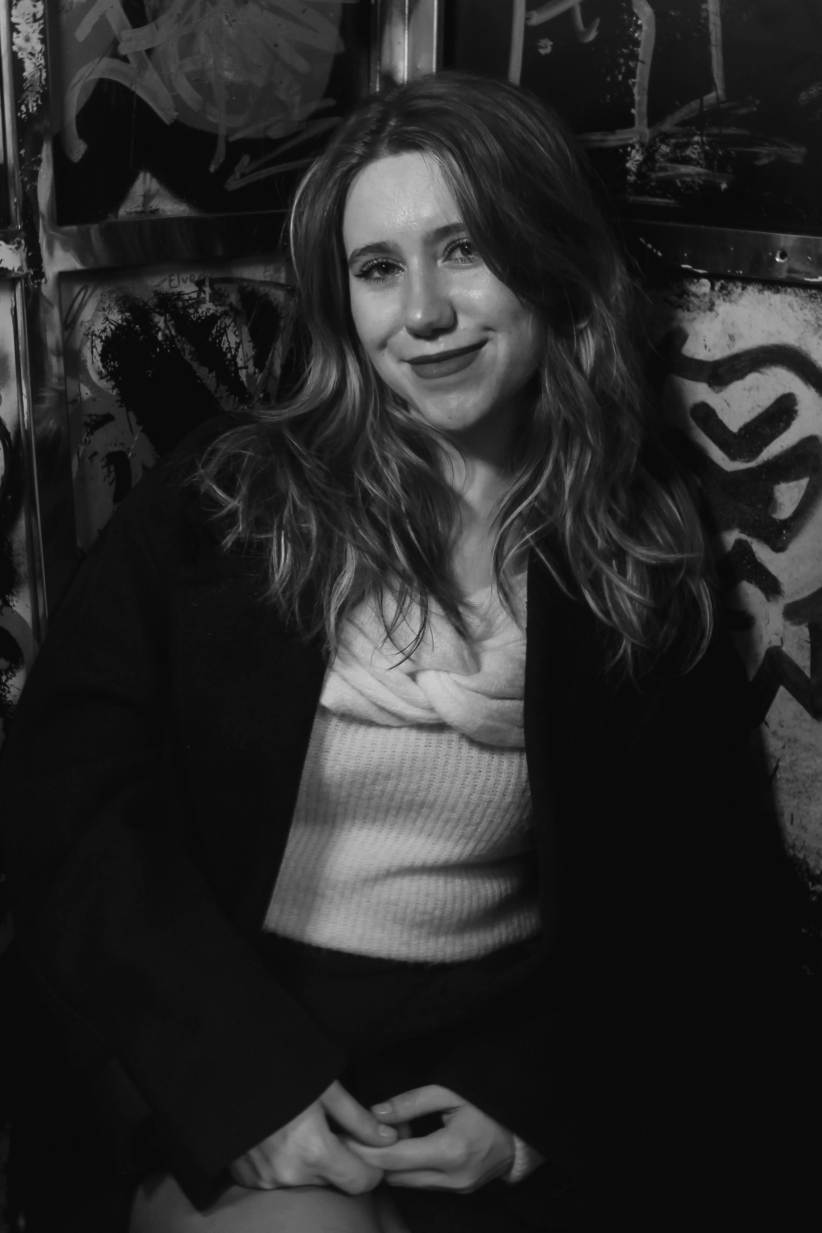 A young adult woman poses in a moody, black and white graffiti-filled environment.