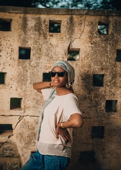 Fashionable woman in sunglasses posing confidently by a rustic stone wall outdoors.