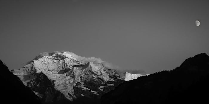 Black and white photo of a snow-capped mountain with the moon in the sky.