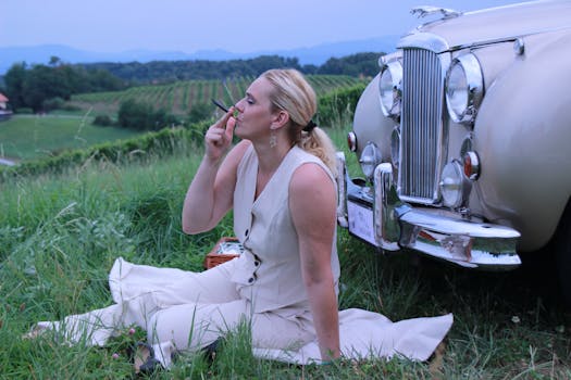 A stylish woman in white enjoys a smoke by a vintage car in a serene vineyard setting.