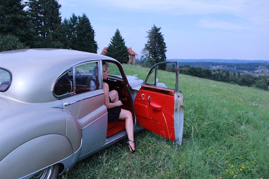 A woman in a black dress sits in a vintage car on a scenic hillside, enjoying the summer view.