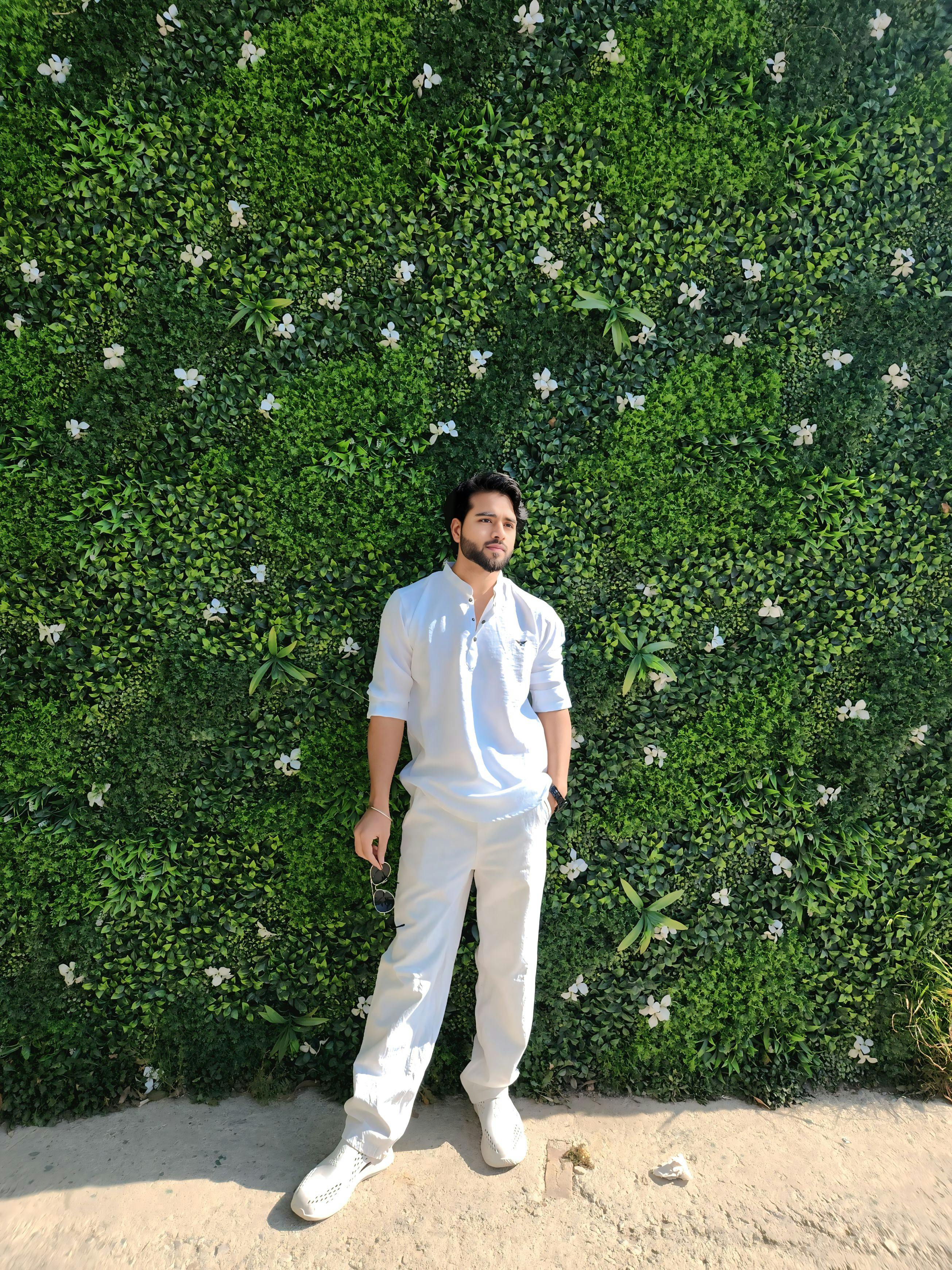 A man stands against a lush green wall, wearing a white outfit.