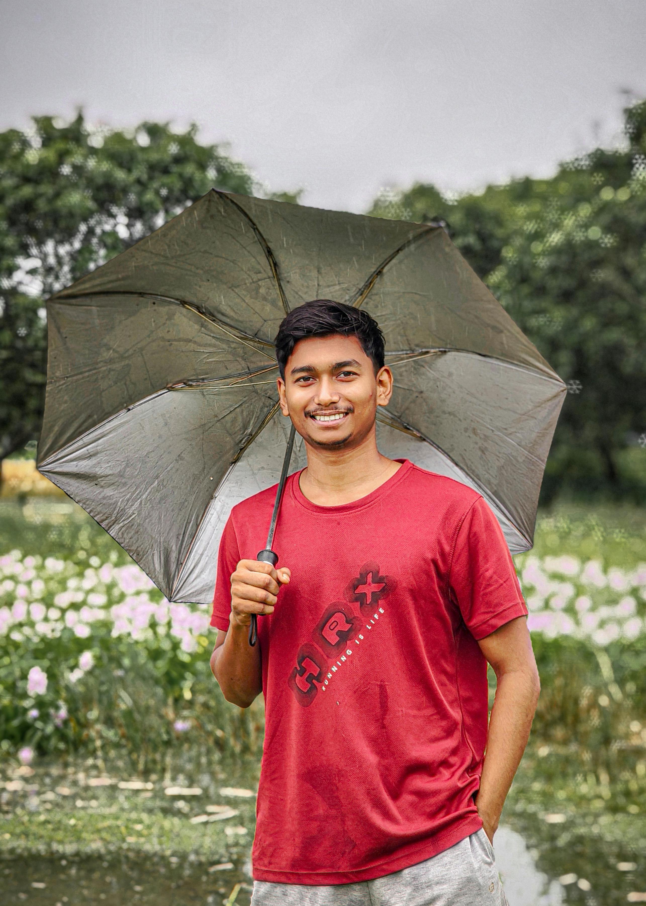 Young man in red shirt holds an umbrella, smiling amidst a blooming field in a rainy outdoor setting.