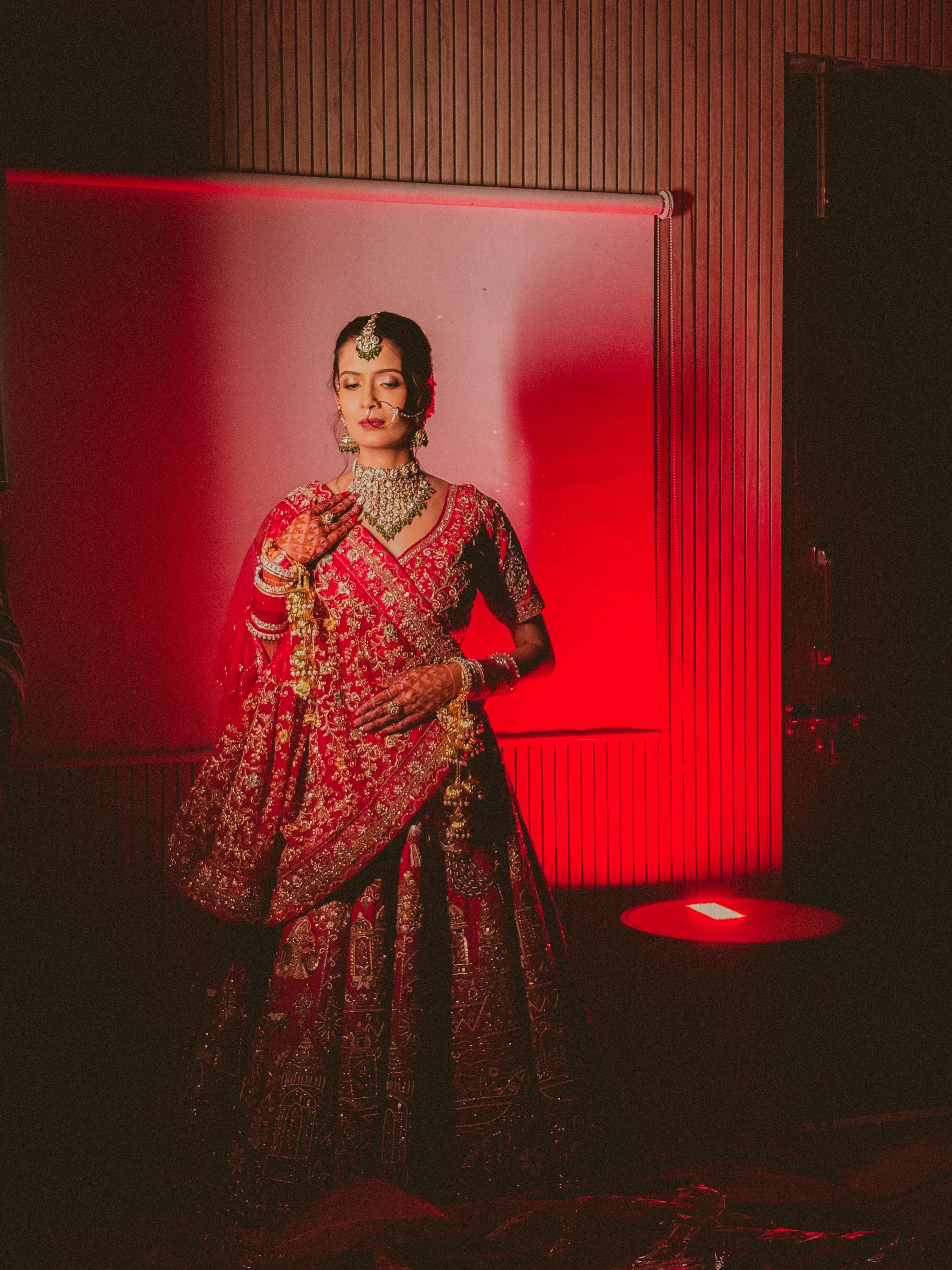Stunning Indian bride in red lehenga stands gracefully in dramatic lighting.