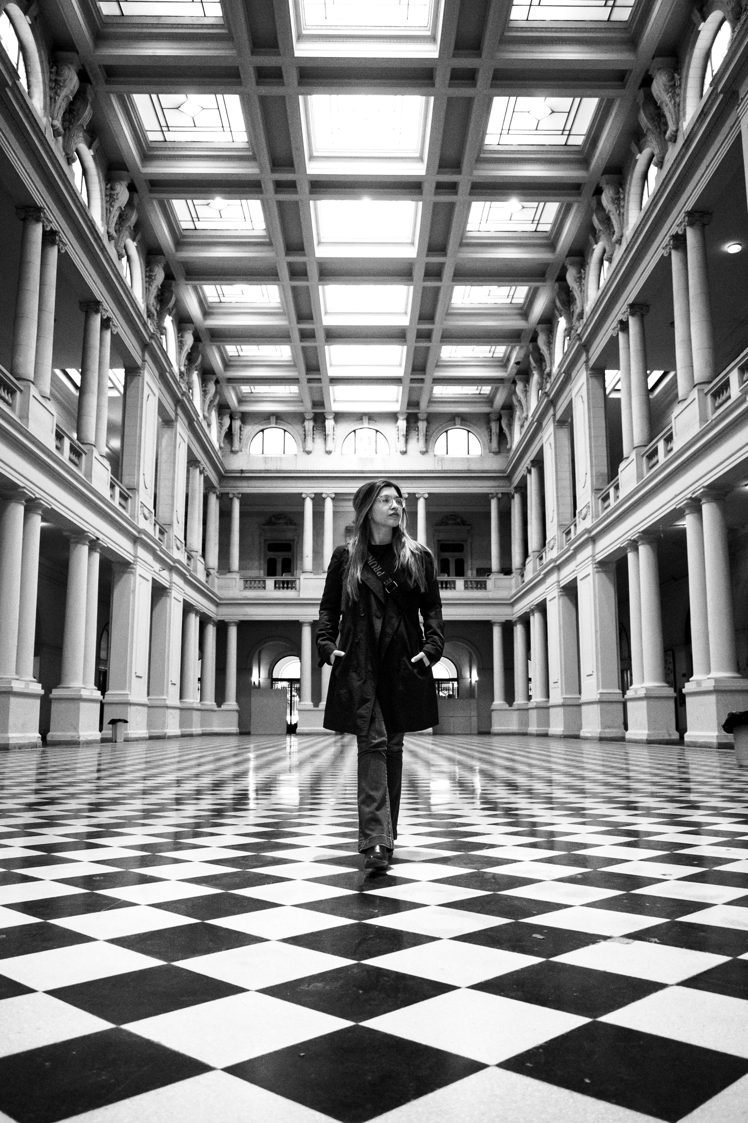 Black and white portrait of a woman walking in a grand hall with checkered flooring.