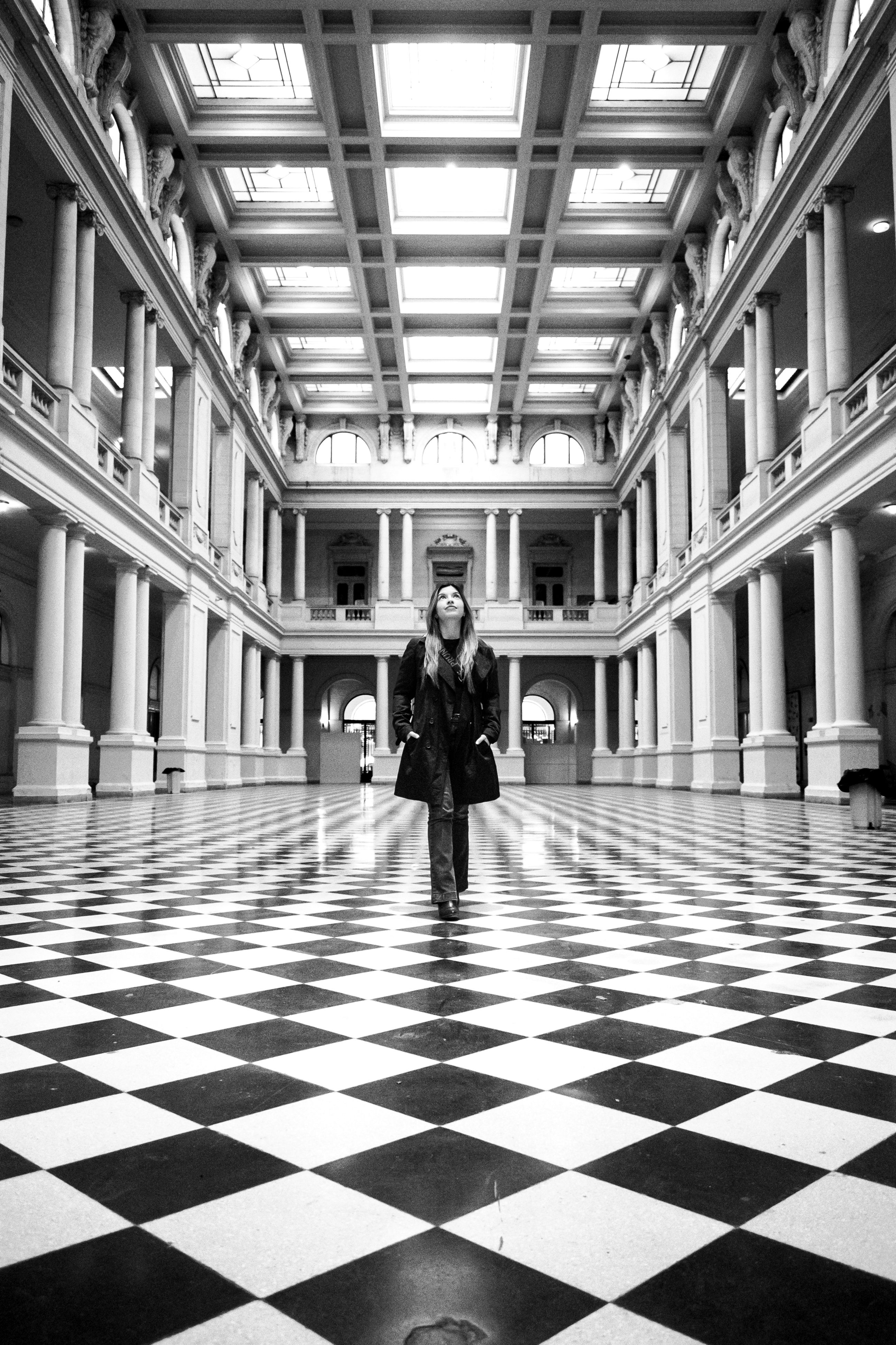A monochrome photo of a person walking in an elegant, grand hallway with checkered floors.