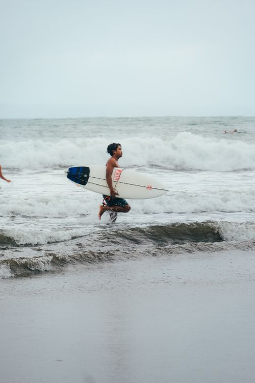 Free A surfer races into the ocean holding a surfboard, ready to catch waves on a cloudy day. Stock Photo