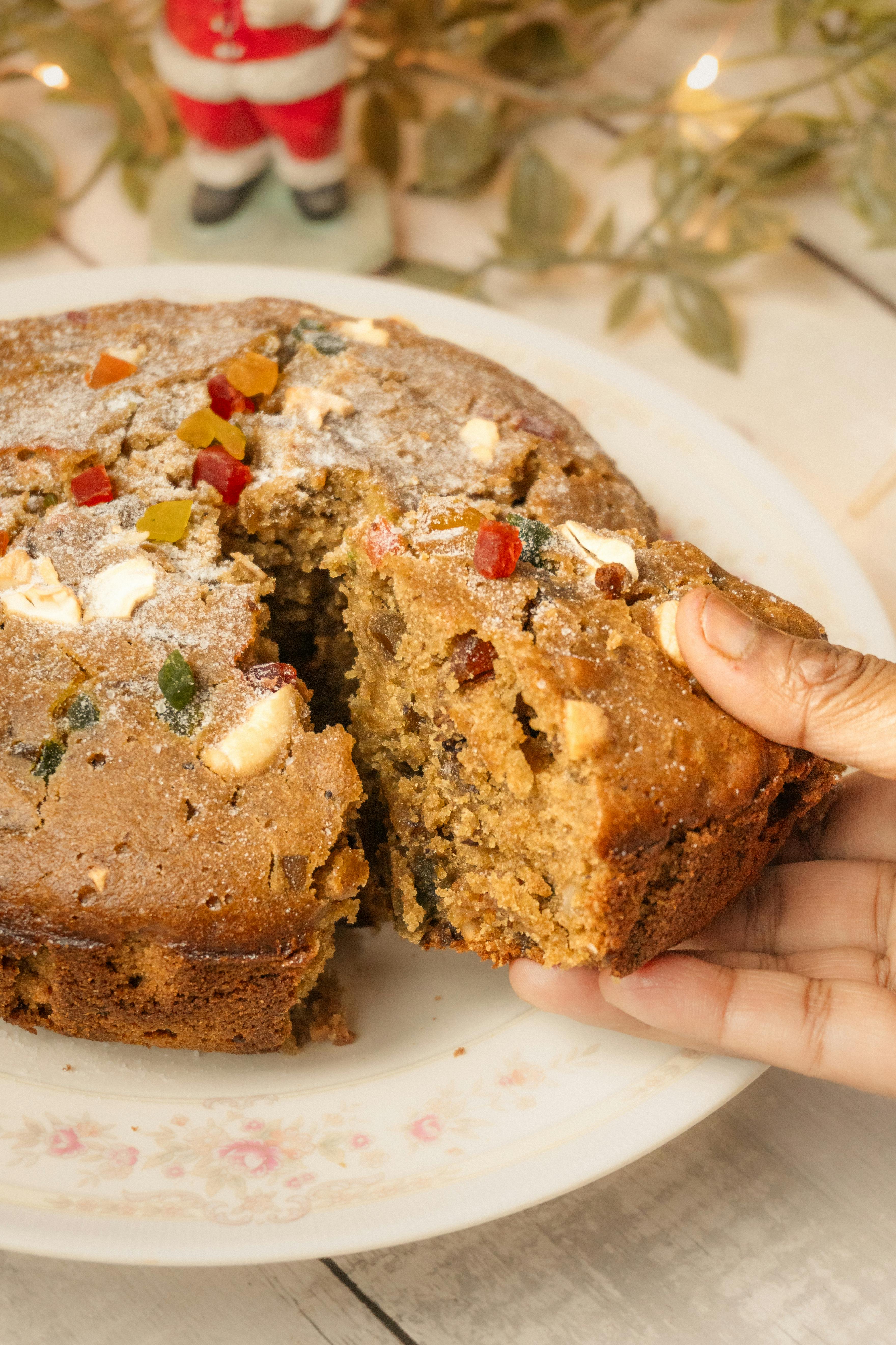 Hand slicing a festive fruit cake with Christmas decorations around.