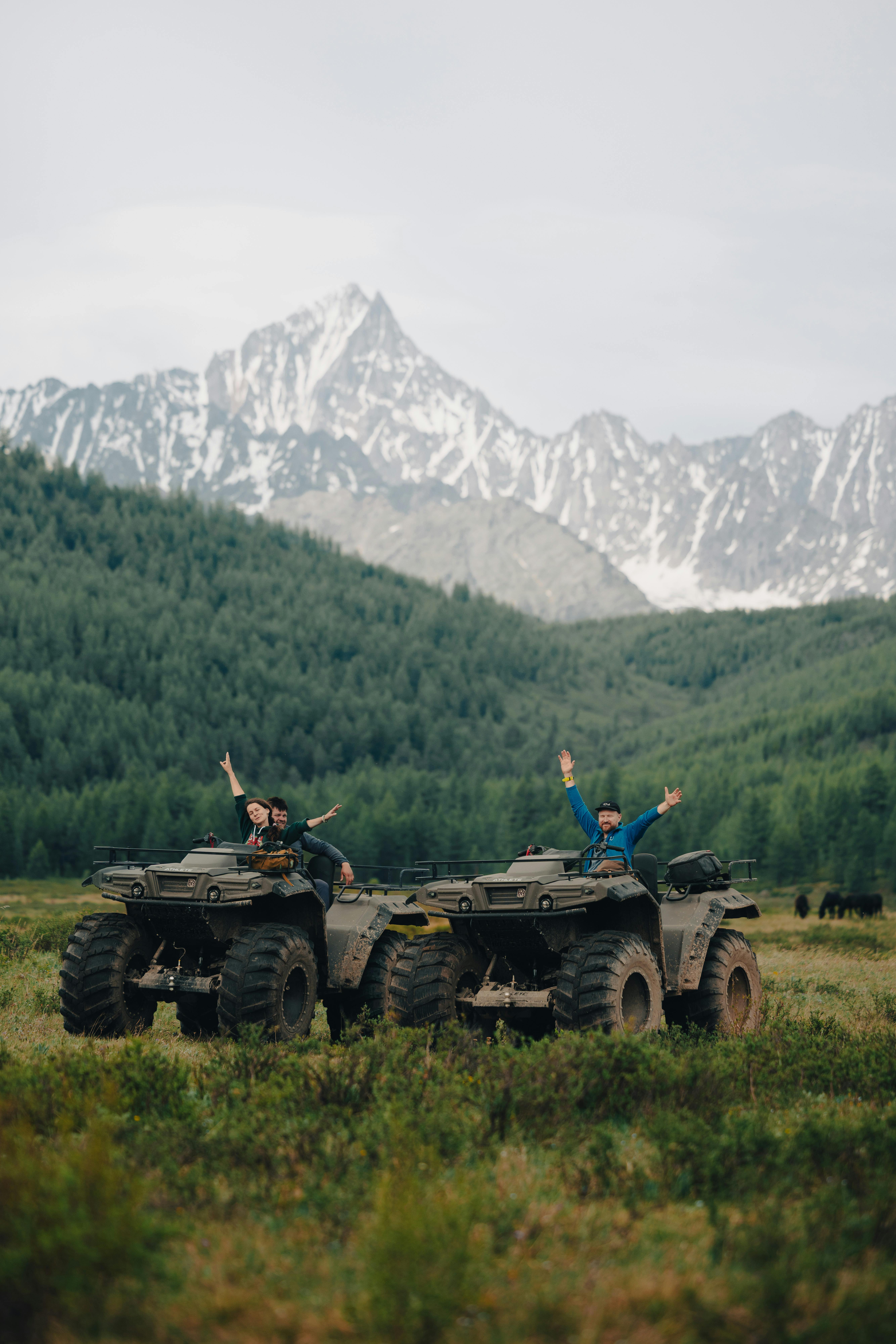 Two people riding ATVs in a mountainous landscape with lush greenery and snow-capped peaks, showcasing outdoor adventure.