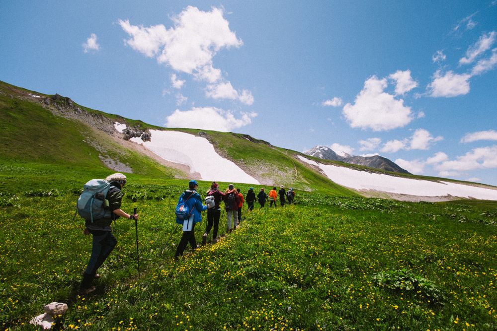 Șir de drumeți cu rucsacuri traversând o pajiște alpină, cu petice de zăpadă