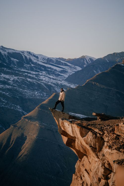 Free A lone person stands on a cliff edge, gazing at the vast, snowy mountain landscape. Stock Photo