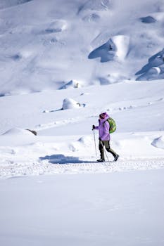 A lone hiker in winter clothing walks through a snowy mountain area with trekking poles.