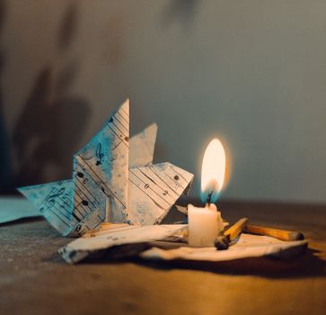 An artistic setting of a paper origami swan made from sheet music, beside a lit candle and matches.