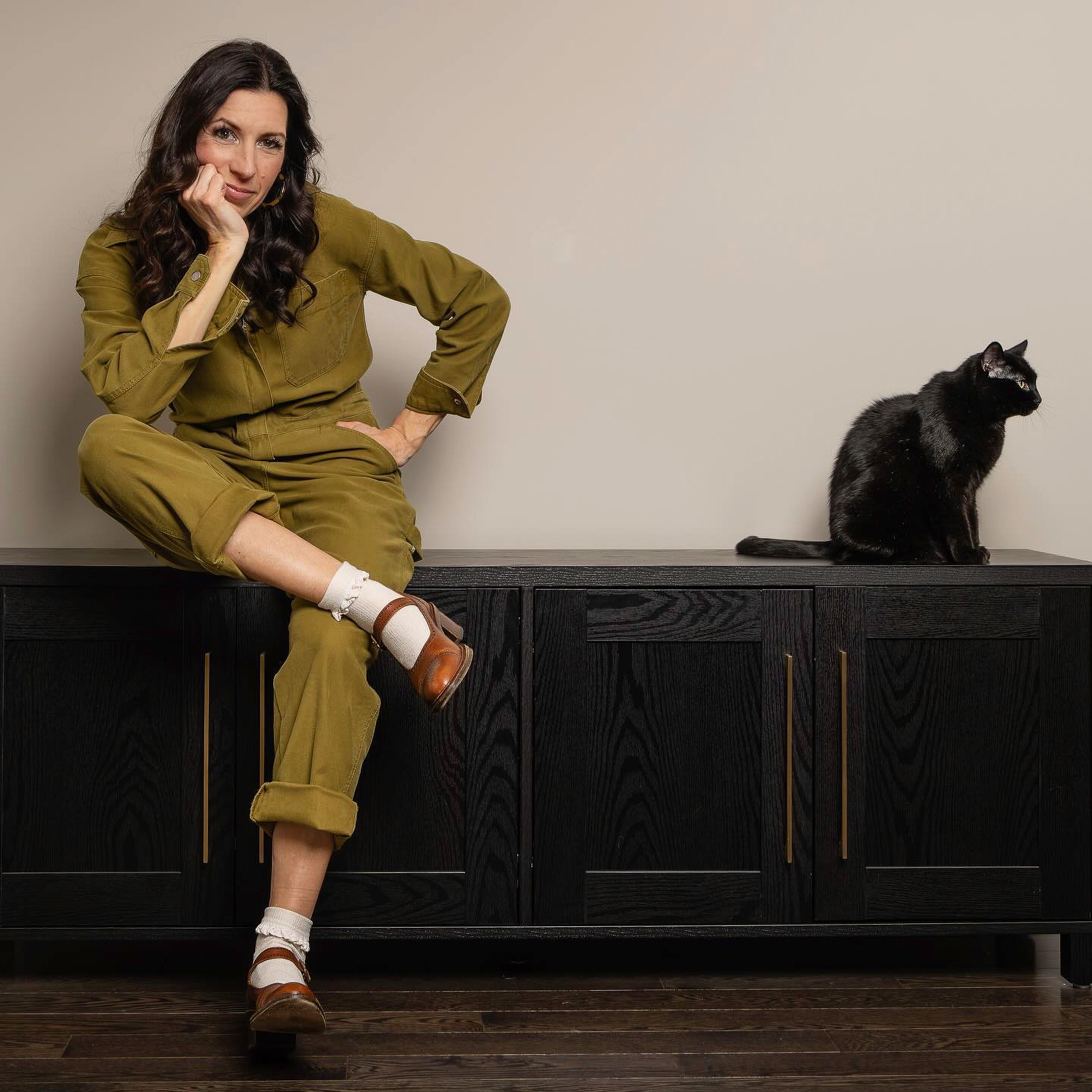 Woman sitting on cabinet with a black cat, both stylishly posed indoors.