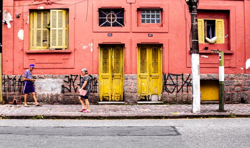 Free Two seniors walk past a vibrant pink building with yellow doors and graffiti. Stock Photo