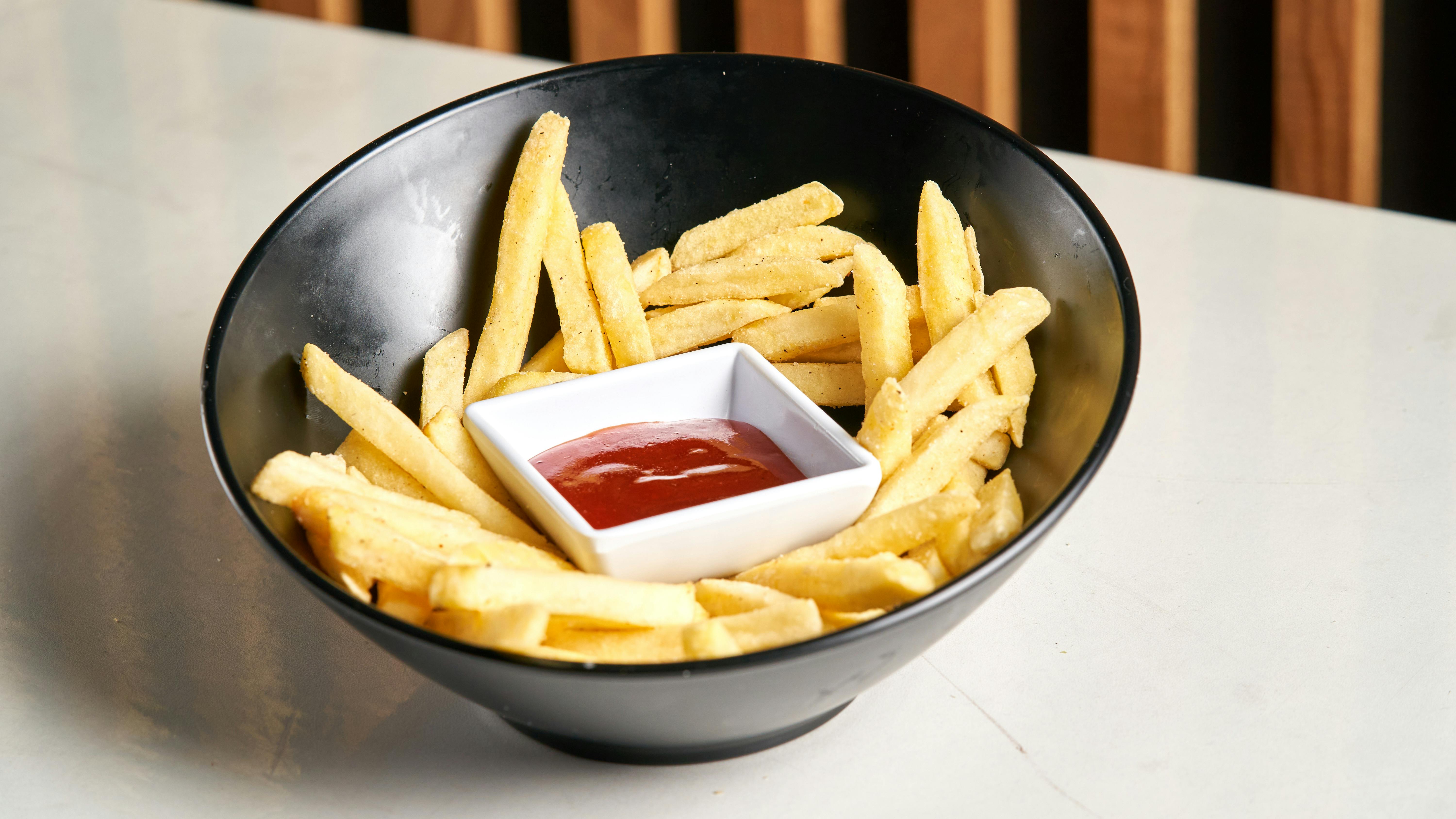 Crispy French fries served with ketchup in a stylish black bowl on a white surface.