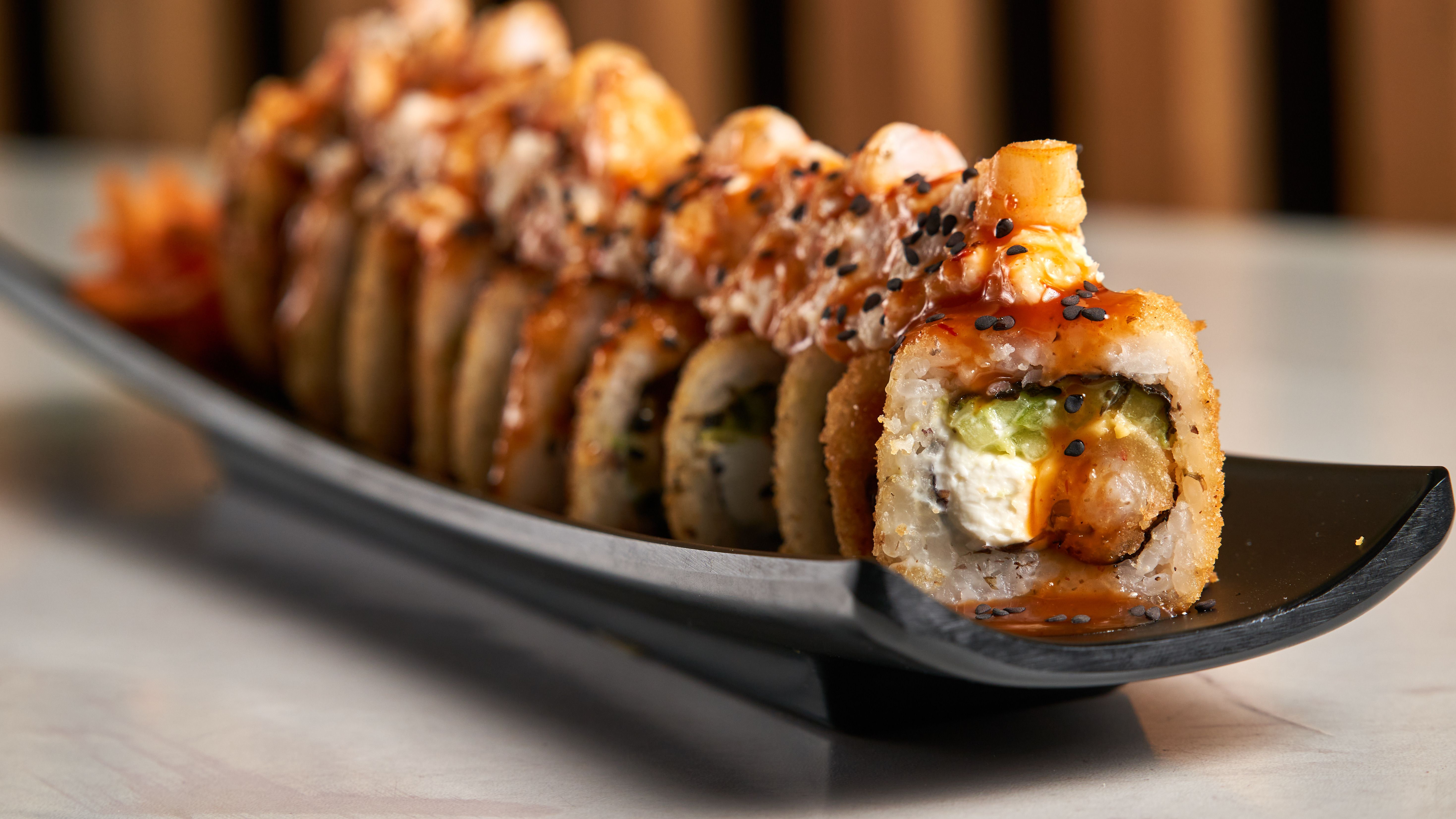 Close-up of tempura sushi roll with vibrant sauce and sesame seeds on black plate.