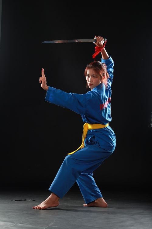 Free Female martial artist in blue gi striking a powerful pose with a sword against a dark background. Stock Photo