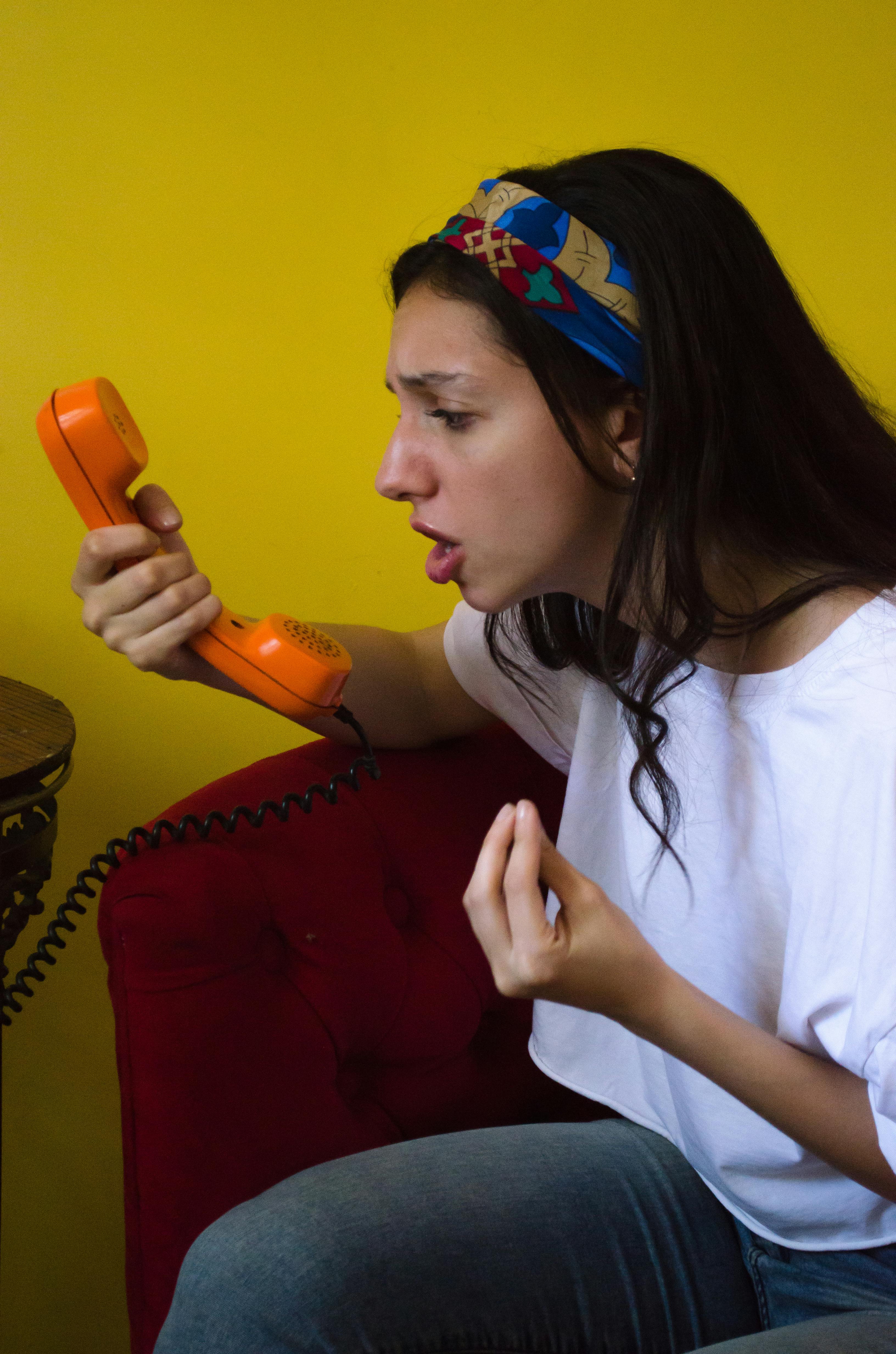 Miễn phí Người phụ nữ trẻ đội băng đô nhiều màu sắc đang tranh luận trên chiếc điện thoại màu cam cổ điển trên bức tường màu vàng sáng. Ảnh lưu trữ