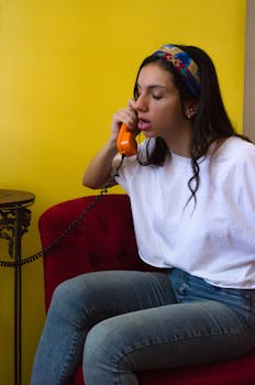 Young woman in casual attire talking on an orange retro phone while sitting indoors.