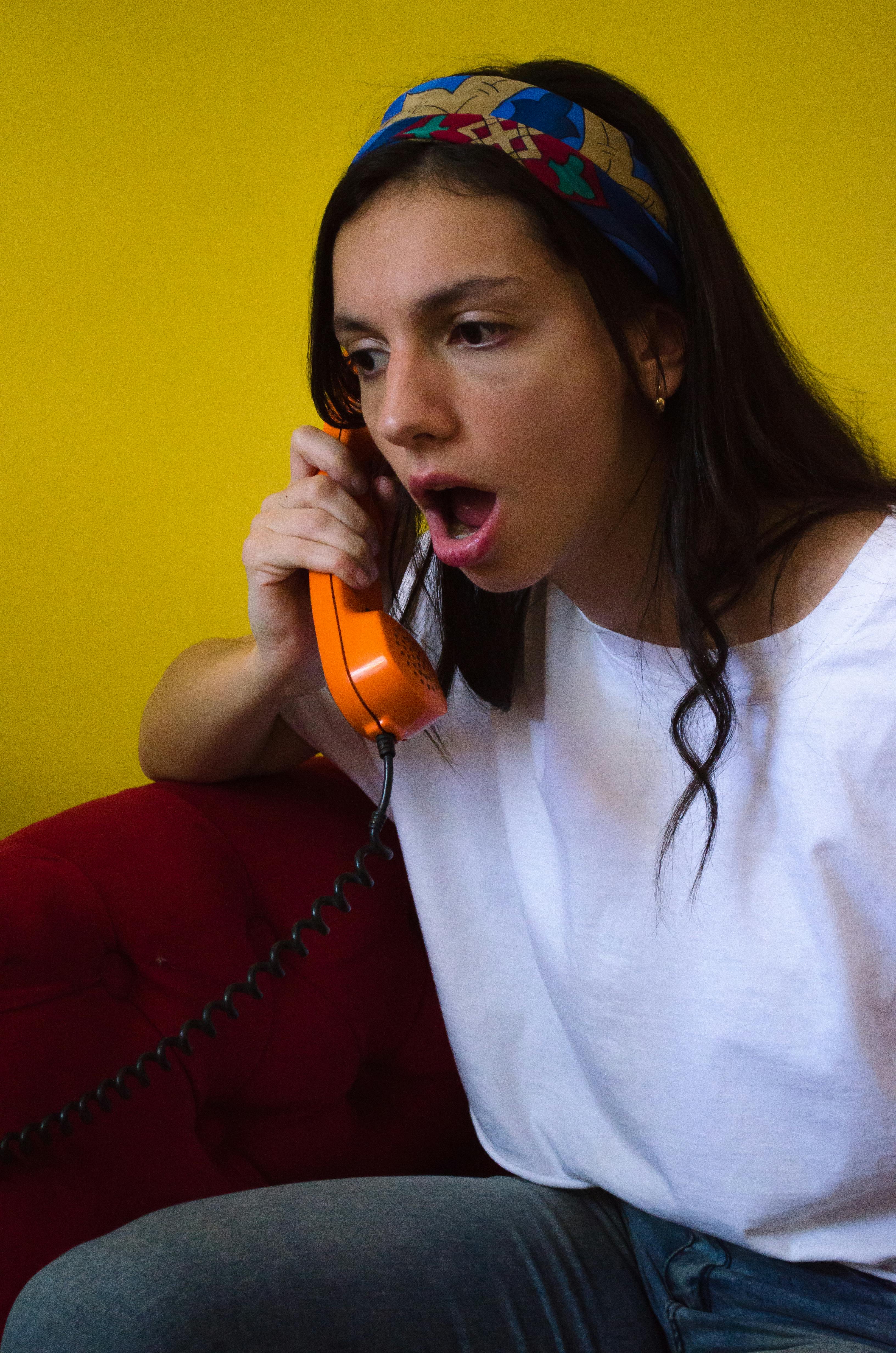 grátis Mulher expressando surpresa durante uma ligação telefônica com um aparelho retrô laranja, contra um vibrante fundo amarelo. Foto profissional