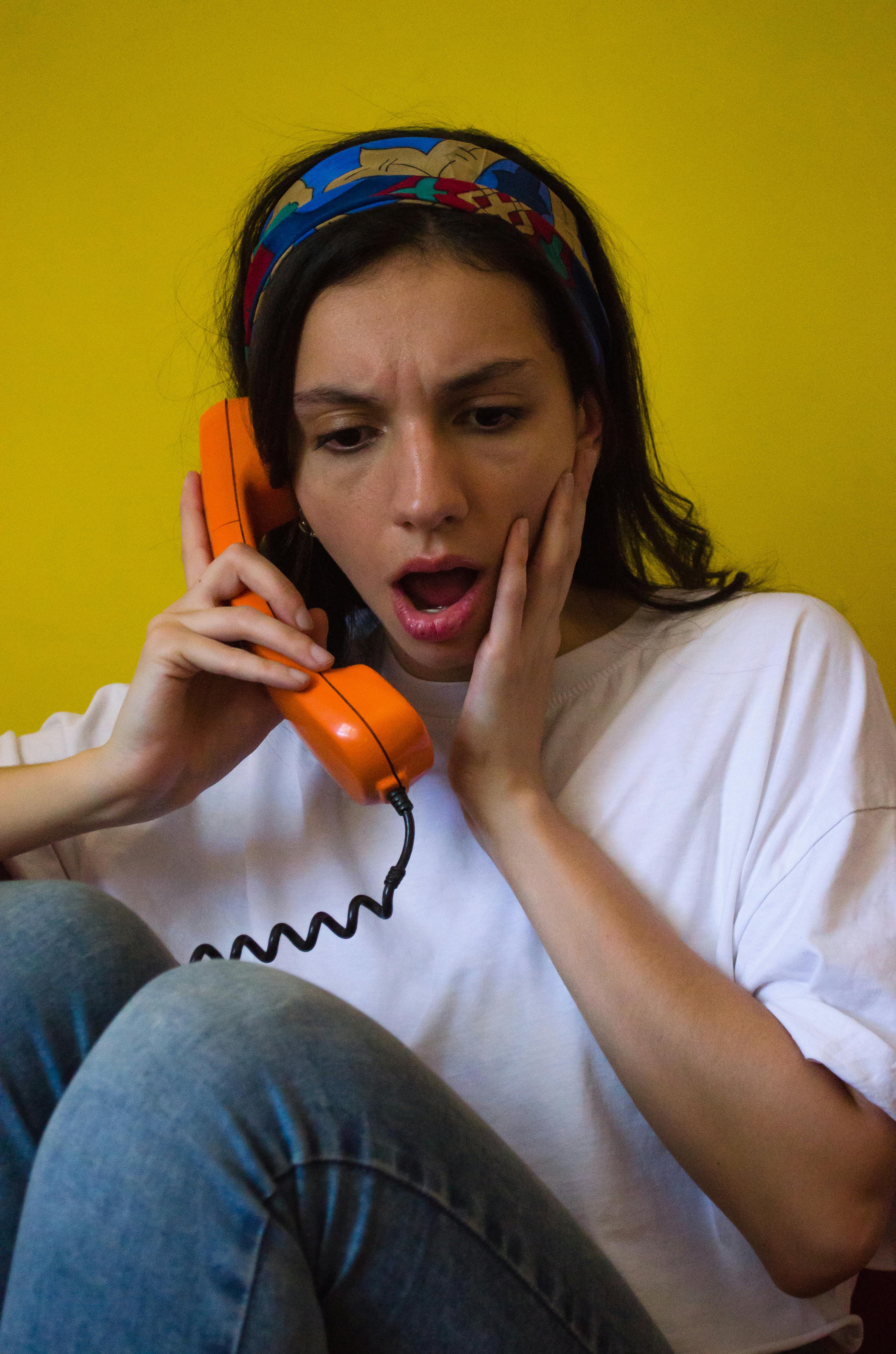 Kostenlos Frau mit überraschtem Gesichtsausdruck telefoniert mit einem altmodischen orangefarbenen Telefon vor gelbem Hintergrund. Stock-Foto