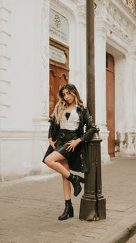 Stylish woman in leather jacket poses by a vintage lamppost in a historical urban setting.