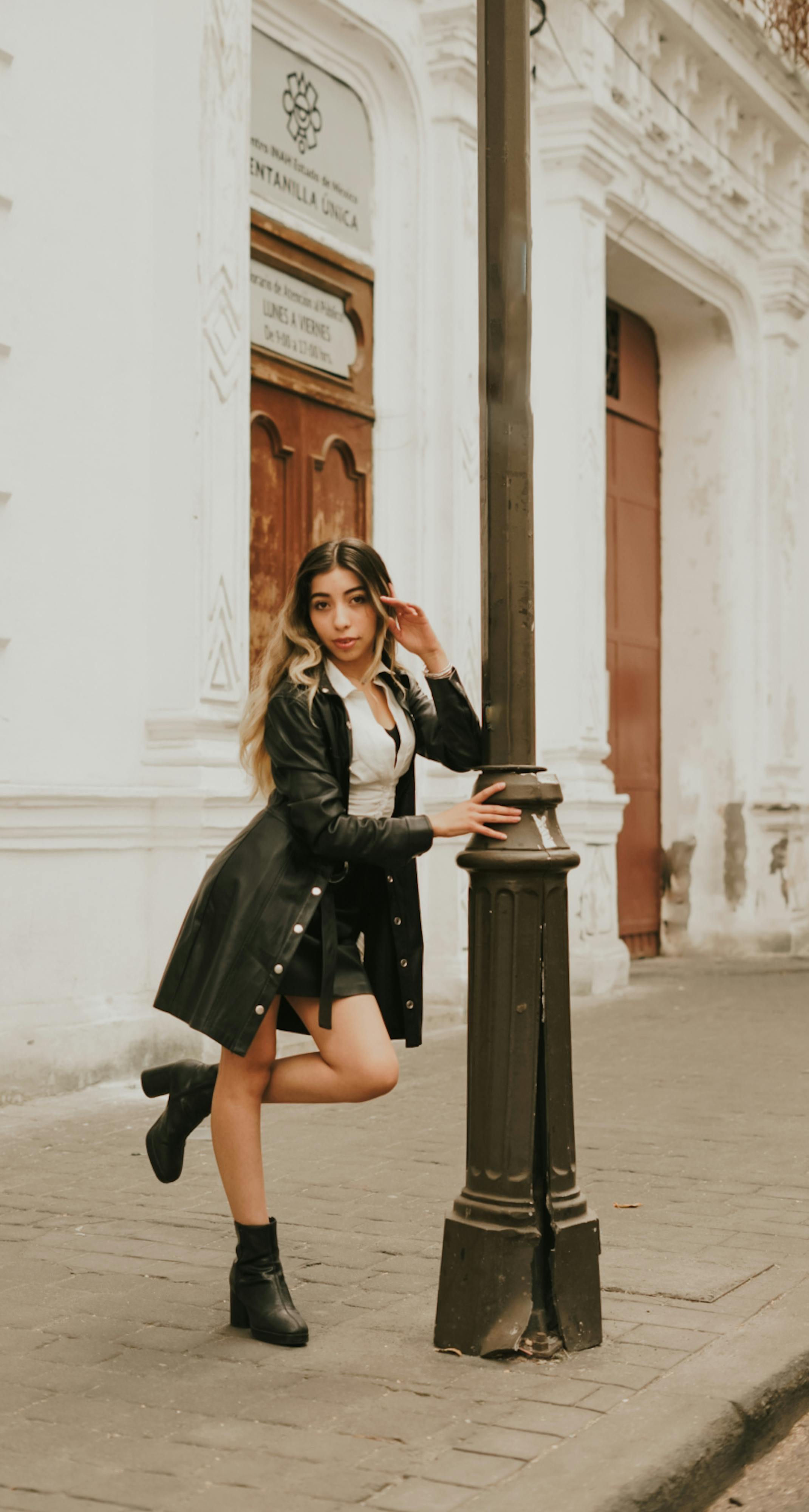 Stylish woman in black leather coat poses confidently by a street lamp in a city setting.