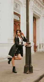 Stylish woman in black leather coat poses confidently by a street lamp in a city setting.