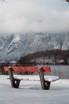A peaceful winter scene with a rustic bench covered in snow, overlooking a tranquil lake and foggy mountains.