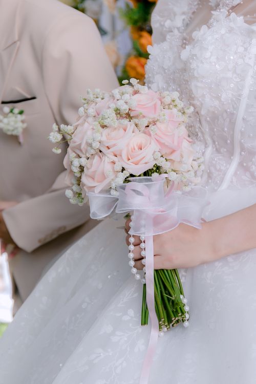 Free Close-up of a bride holding a pink rose bouquet with pearls and lace in a soft, romantic wedding setting. Stock Photo