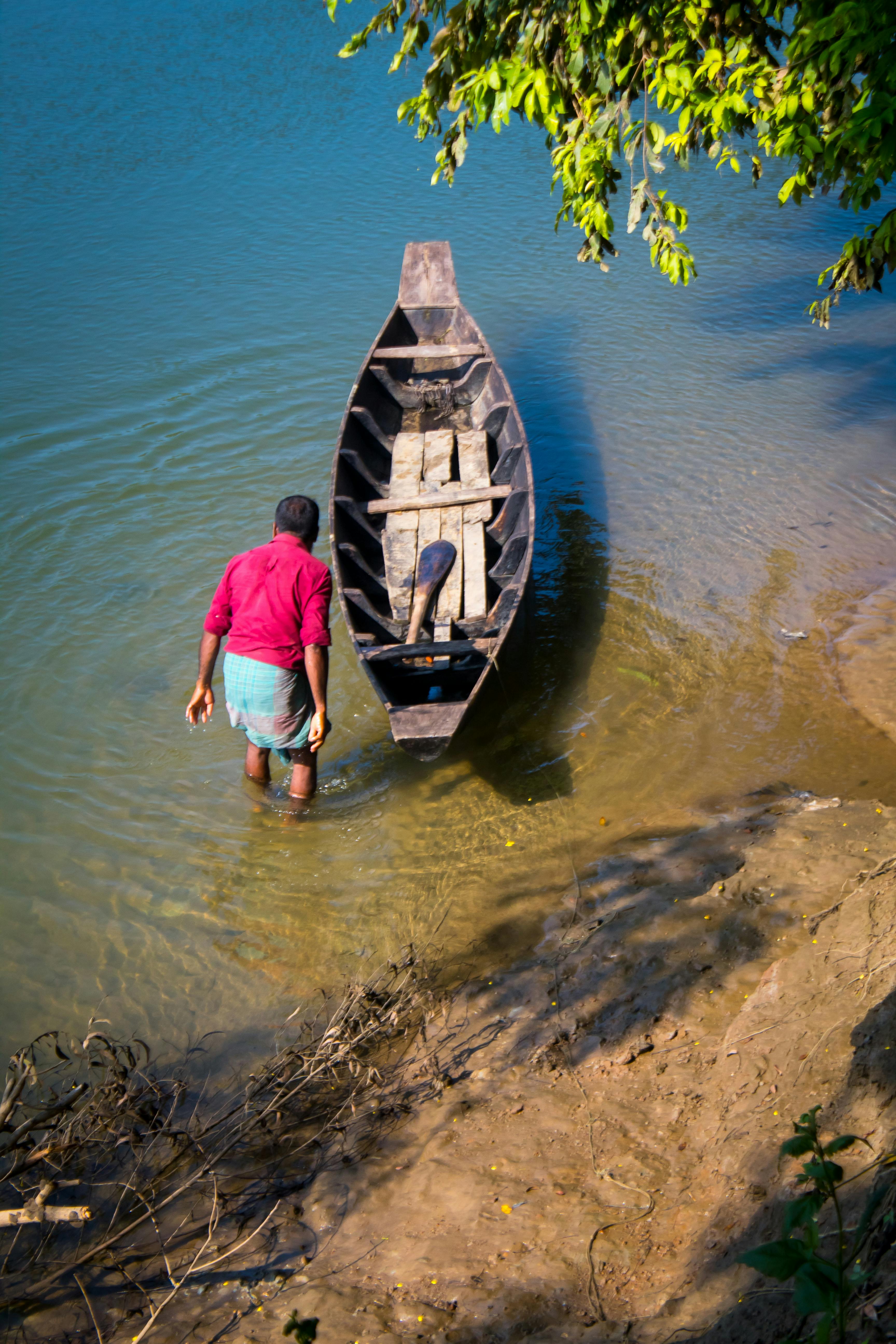 A man stands beside a wooden boat on a serene riverbank in Rangamati, Bangladesh.