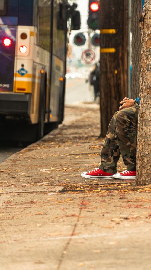 Free A person in red shoes and camo pants sits beside a tree on a urban street with a bus nearby. Stock Photo