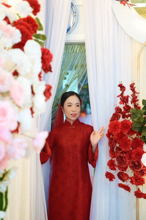 Free Woman in red traditional dress standing amidst vibrant floral decor indoors. Stock Photo